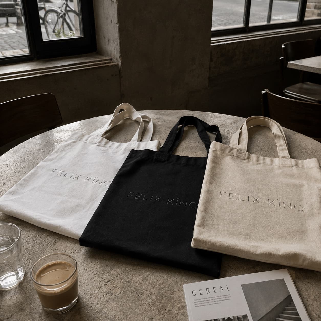 Three canvas tote bags in white, black and natural, each embroidered with the Felix Kïno wordmark, laid out on a café table